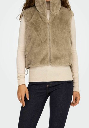 Beige Kunstpelz-Weste mit Reißverschluss, getragen über einem hellbeigen Langarmshirt und dunklen blauen Jeans an einer stehenden Person.