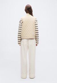 Άτομο που βρίσκεται με την πλάτη στην κάμερα, φοράει μπεζ γιλέκο με ραφές, ριγέ πουκάμισο με μακριά μανίκια και άνετο λευκό παντελόνι μπροστά σε απλό φόντο.