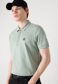 Chemise polo vert clair avec un col, deux boutons et une écusson texturé sur la poitrine. Portée avec un jean noir slim fit et une casquette noire.