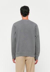 En mann som vender bort med en enkel grå langermet sweatshirt og beige bukser mot en hvit bakgrunn.