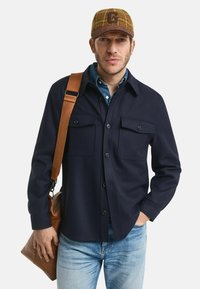 Camicia blu con bottoni, dotata di due tasche sul petto, indossata sopra una camicia di denim, abbinata a jeans sbiaditi e una borsa a tracolla in pelle marrone.