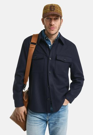 Marineblauwe button-up shirt met twee borstzakken, gedragen over een denim shirt, gecombineerd met vervaagde jeans en een bruine leren schoudertas.