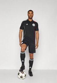 Black soccer uniform with red and white stripes, short sleeves, and shorts. Player stands on a soccer ball, wearing black cleats and striped socks.