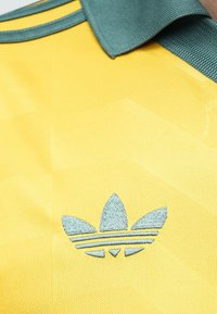 Geltonos spalvos audinys su siuvinėta mėlyna Adidas trefoil logotipu ir dalimi mėlynos apykaklės ant tekstūruoto marškinio.