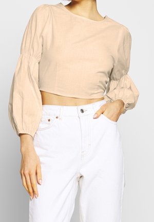 Mujer con una blusa corta beige de mangas largas abullonadas y vaqueros blancos de talle alto, con una mano en el bolsillo.