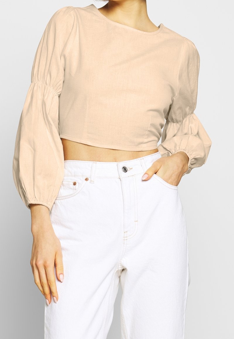Femme portant un chemisier court beige à manches longues bouffantes et un jean blanc taille haute, une main dans la poche.