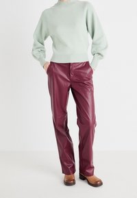 Personne portant un pull vert clair, un pantalon en cuir synthétique bordeaux et des bottines marron, debout les mains dans les poches sur un fond blanc.
