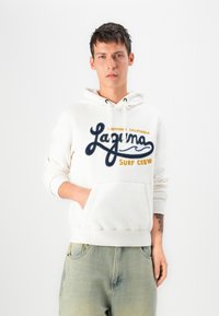Felpa bianca con ricamo blu navy e giallo "Laguna Surf Crew", dotata di tasca frontale e polsini a coste. Indossata con jeans verdi scoloriti.