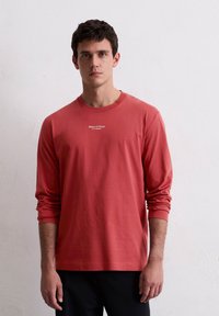 Langarm, roestkleurig T-shirt van zachte katoen. Bevat een ronde halslijn en het merklogo gedrukt op de borst.