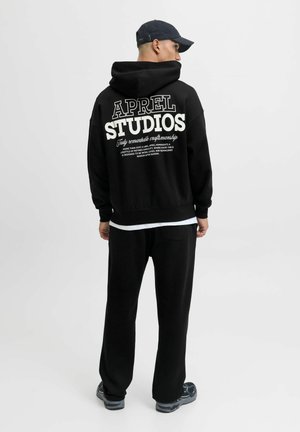 Schwarzer Hoodie mit dem Schriftzug "APREL STUDIOS" in Weiß auf dem Rücken, kombiniert mit schwarzen Sweatpants. Hat eine entspannte Passform und eine weiche Textur.