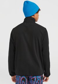Giacca nera con colletto, realizzata in materiale liscio. In vista anche un berretto blu a maglia e un pantalone a fantasia multicolore.