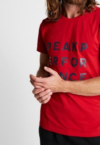 Camiseta de algodón roja con mangas cortas, que presenta letras negras en negrita que dicen "PEAKPERFORMANCE" en la parte frontal.