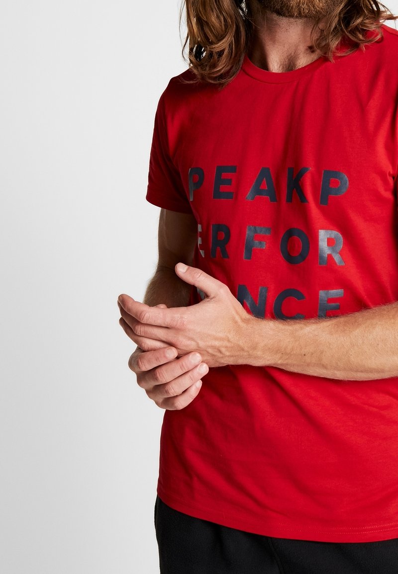 Camiseta de algodón roja con mangas cortas, que presenta letras negras en negrita que dicen "PEAKPERFORMANCE" en la parte frontal.