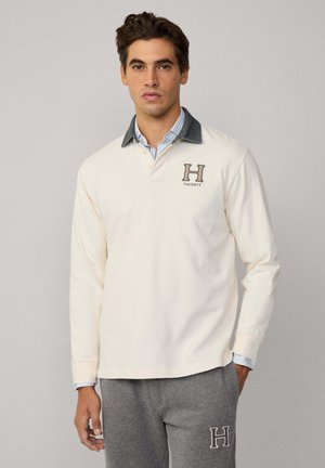 Polo a maniche lunghe color crema con colletto scuro, caratterizzato da un logo "H" e dal testo "HACKETT" in colori a contrasto; indossato con pantaloni della tuta grigi.