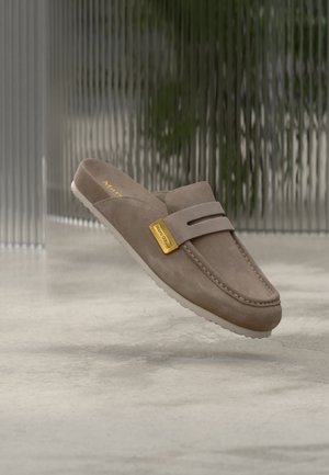 Mocassin en daim taupe à enfiler avec détail de boucle dorée, semelle blanche et coutures visibles, présenté sur un fond neutre flou.