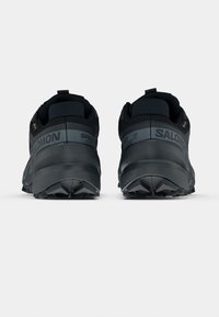 Čierne atletické topánky s textúrovaným materiálom, s vystuženou pätou a výrazným vzorom na podrážke. Na bokoch sú viditeľné logo a názov modelu.
