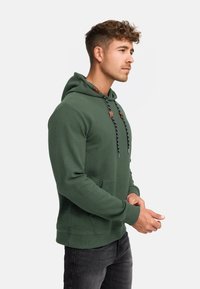 Grøn hættebluse i strukturstof med en forlommer, sorte og hvide snore og brune detaljer på hætten. Casual fit.