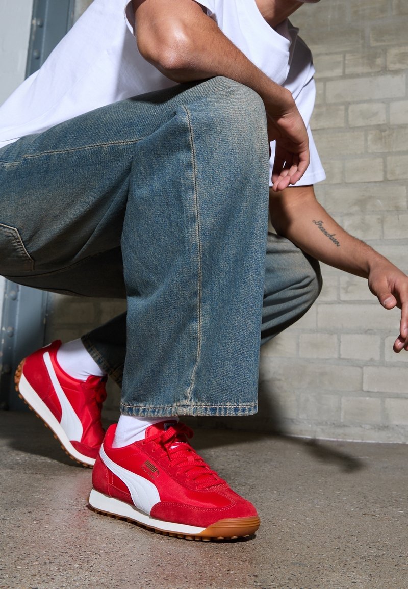 Personne accroupie portant des baskets Puma rouges et blanches, un jean bleu clair, des chaussettes blanches et un t-shirt blanc devant un mur de briques grises.