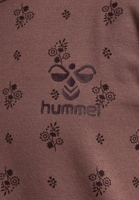 Brun stof med et struktureret mønster af mørke blomster og "hummel"-logoet prentet i sort, centreret og symmetrisk over materialet.