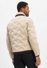 Veste matelassée beige avec col montant, accents noirs, tissu texturé et poignets côtelés. Coutures plates et coupe décontractée.