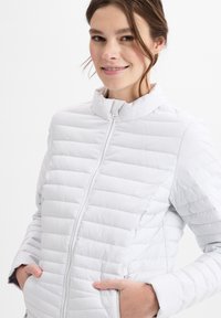 Hellegraue gepolsterte Jacke mit hohem Kragen, Frontreißverschluss und Seitentaschen. Verfügt über einen strukturierten Stoff mit horizontalem Steppmuster.