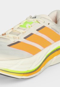 Chaussure de course blanche avec trois bandes orange, des touches jaune néon, des lacets blancs et une semelle épaisse et rembourrée sur fond gris clair.