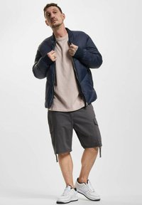 Marineblauer Pufferjacke mit gerippten Bündchen, beigefarbenem Text hemd, grauen Cargo-Shorts und weißen Sneakern mit schwarzen Akzenten.