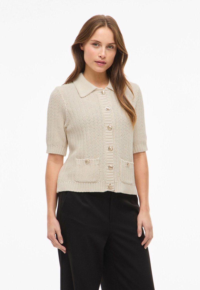 Beżowy kardigan krótki rękaw, z teksturowanym wzorem dzianiny, ostrym kołnierzem i guzikami z przodu. Posiada dwie przednie kieszenie z akcentami guzikowymi.