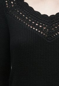 Sweater preto em malha com gola de croché em forma de concha e tecido texturizado. Vista em close-up revela detalhes intrincados e padrões de pontos.