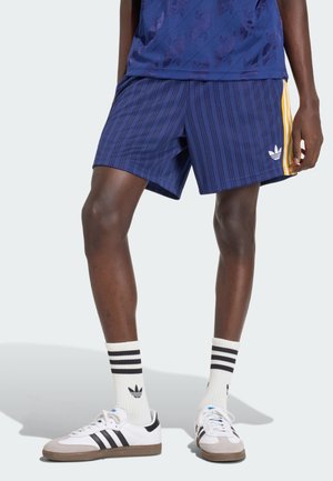 Persoon draagt blauwe sportshorts met gele zijstrepen, witte Adidas-sokken met zwarte strepen en witte sneakers met zwarte strepen.