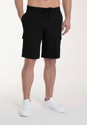 Uomo che indossa pantaloncini cargo neri al ginocchio con tasche laterali e scarpe da ginnastica bianche, in piedi con le mani rilassate lungo i fianchi.