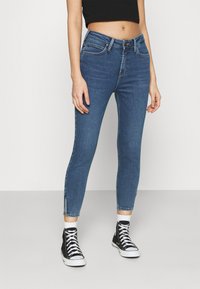 Jeans de mezclilla azul de corte skinny con una cintura de altura media, que cuentan con bolsillos frontales y dobladillos con cremallera, combinados con zapatillas negras de caña alta.