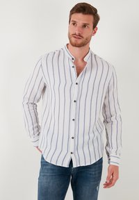 Camisa de botones hecha de una tela ligera, con rayas verticales azules y blancas, cuello de pie y botones oscuros, combinada con jeans azules.