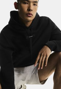 Zwarte hoodie met drawstrings, voorzien van een eenvoudig logo label, gecombineerd met lichtgrijze shorts. Glad textuur, urbane casual stijl.