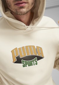 Felpa color crema con una grande grafica "PUMA SPORTS" in oro, nero e verde. Presenta un cappuccio con cordini e una texture morbida.
