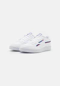 Ténis brancos feitos de pele lisa, com um design de cano baixo, fecho com atacadores e detalhes em vermelho e azul nas laterais e na língua.