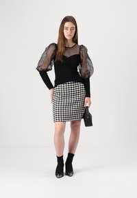 Zwarte top met doorzichtige pofmouwen en kraalaccenten, gecombineerd met een getailleerde houndstooth rok. Model houdt een kleine zwarte handtas vast.