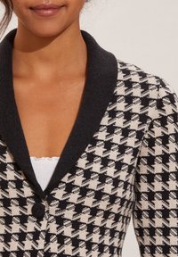 Musta-valkoinen houndstooth-kuvioinen neuletakki, jossa on tumma kaulus, teksturoitu neulekangas, yksittäinen napitus ja vartalonmyötäinen muotoilu.