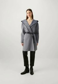 Žena oblečená v šedej zavinovacej bundě s opaskom a vzorovaným širokým golierom, čiernych skrátených nohaviciach a čiernych členkových topánkach stojaca na svetlom pozadí.