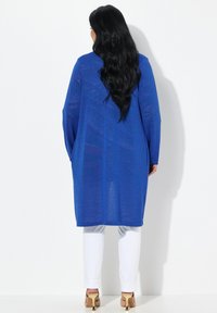 Lang, blauw, lichtgewicht cardigan met een subtiele glans, voorzien van brede mouwen en een losse fit, gecombineerd met witte broeken en beige hakken.
