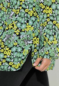 Blusa floreale con base nera decorata da fiori verdi e gialli vivaci, tessuto con texture e spacco laterale per una maggiore libertà di movimento.