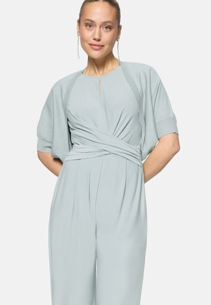 Žena oblečená v svetlomodrom zavinovacom overale s krátkym kardigánom a dlhými náušnicami, stojaca s rukami za chrbtom.