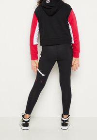 Bambino che indossa leggings neri con un logo bianco e rosso sulla gamba destra, felpa nera con maniche rosse e bianche e sneakers bianche e nere.