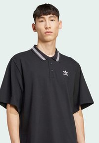 Black polo shirt made of textured fabric with a three-button placket and a striped collar. Features small white adidas logo on the chest.