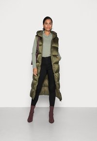 Gilet lungo imbottito verde oliva con cappuccio, indossato sopra una maglietta verde chiaro, abbinato a pantaloni neri attillati e stivaletti larghi bordeaux.