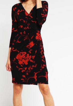Kobieta w czarnej, sięgającej kolan sukience z czerwonymi kwiatowymi wzorami i rękawami do połowy ramienia, pozująca z jedną ręką przy szyi.