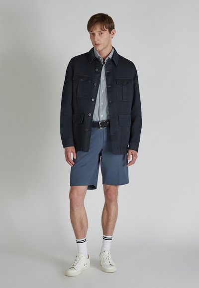 Veste en coton navy avec poches avant, accompagnée d'une chemise à rayures légères, de shorts gris et de baskets blanches avec des accents et des rayures noires.