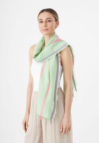 Écharpe vert clair avec des motifs zigzag rose et violet, ornée de bords à franges. Portée enroulée sur l'épaule, texture douce.