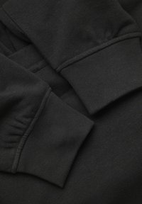 Tessuto in cotone nero per felpa con polsini a costine e texture liscia. La vista ravvicinata mette in evidenza i dettagli delle cuciture e la vestibilità elastica.