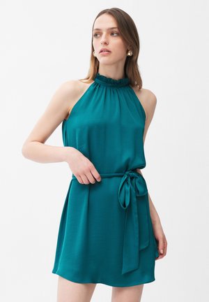 Kobieta w turkusowej sukience na szyję bez rękawów z falbankowanym kołnierzykiem i paskiem w talii, pozująca na białym tle.